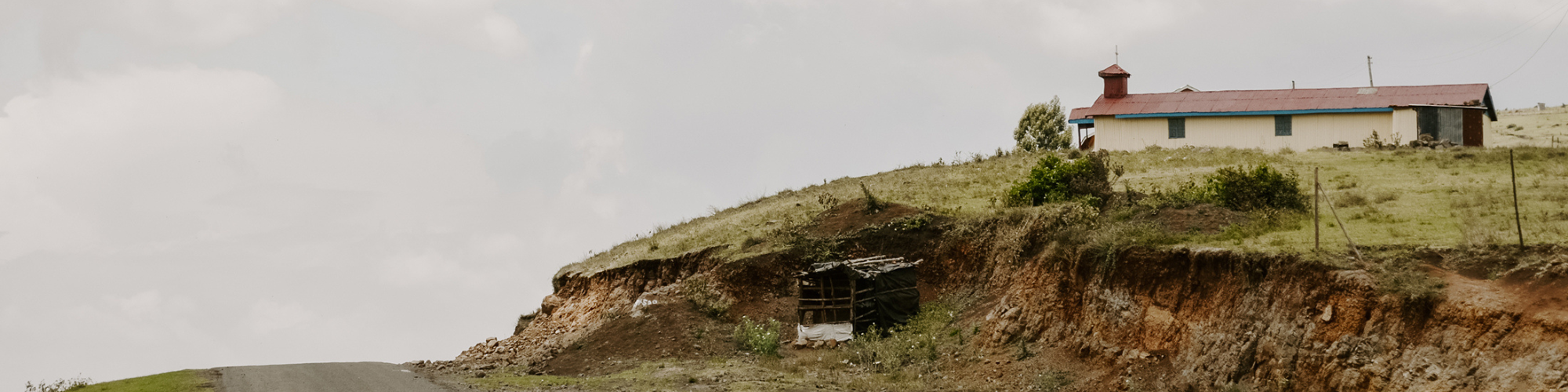 A small, simple building with a red roof, perched on a hill next to a dirt road. The landscape is sparse, with the building standing alone against a backdrop of cloudy skies.