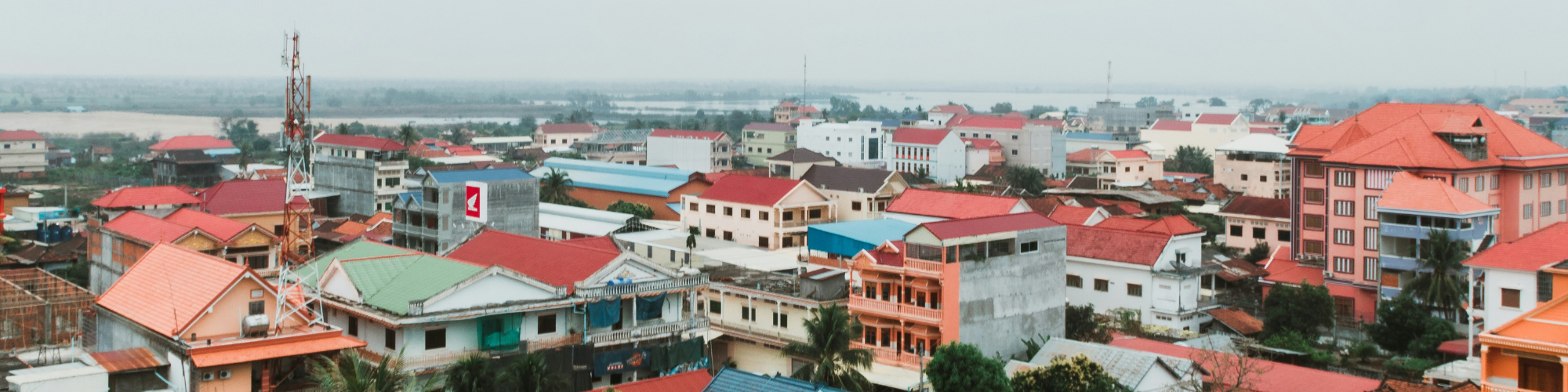 A panoramic view of a town with colorful rooftops in shades of red, orange, green, and blue. The buildings vary in size and style, with some modern and others more traditional. In the background, a river and flat landscape stretch into the horizon under a cloudy sky, giving a peaceful and expansive feel to the scene.