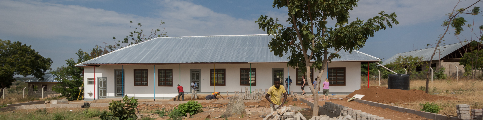 A construction site in progress with workers laying bricks and performing tasks in front of a newly built single-story building with a metal roof, surrounded by a dusty, unfinished landscape and a few trees under a partly cloudy sky.