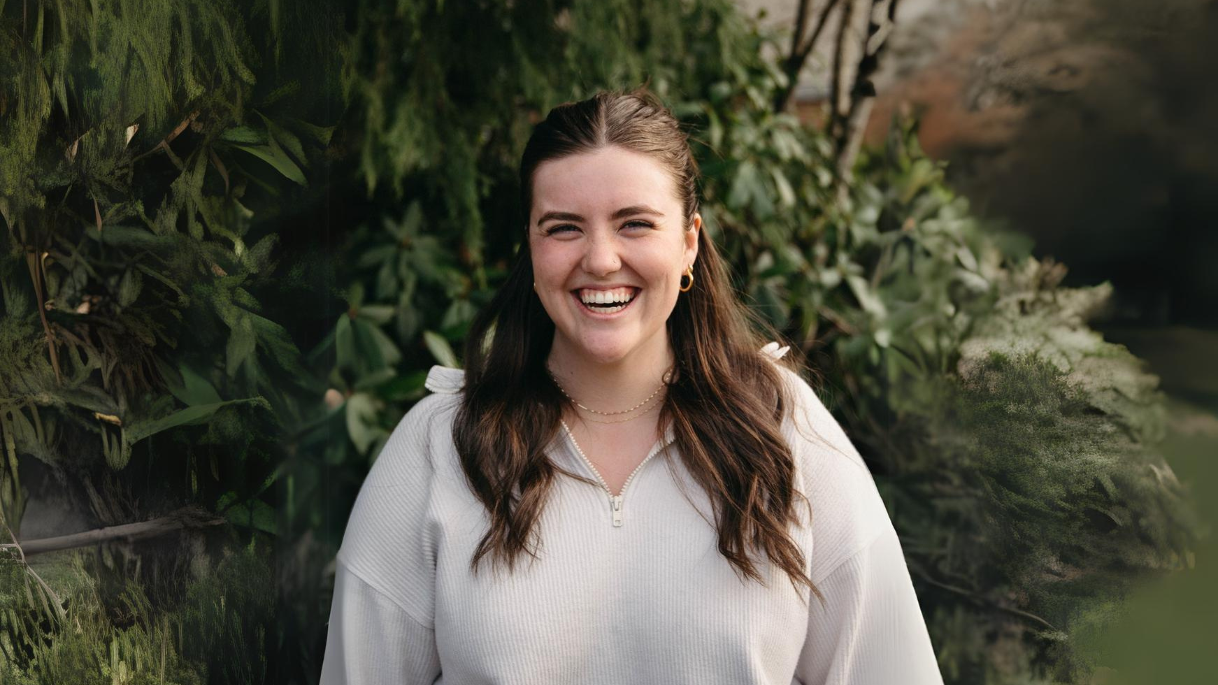  A young woman with long brown hair is smiling brightly while standing outdoors. She is wearing a light-colored zip-up sweater and a delicate necklace. The background features green foliage, creating a natural, pleasant atmosphere.