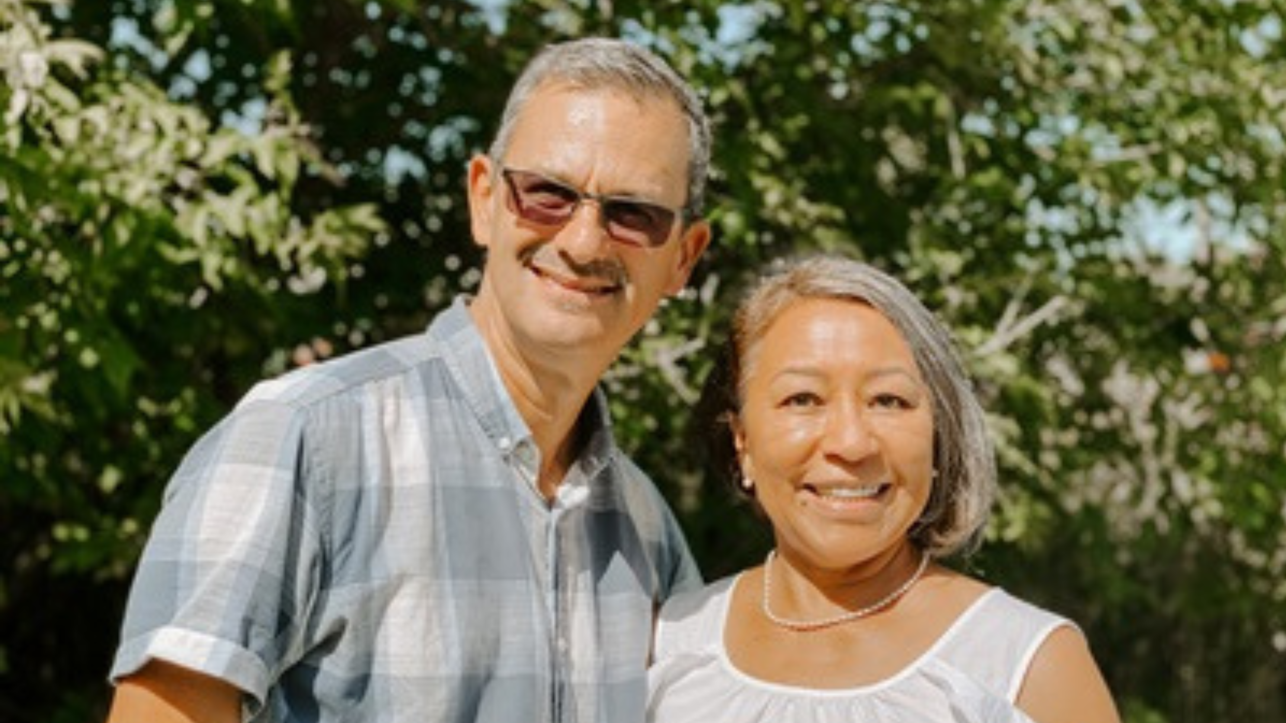 Outdoor portrait of a middle-aged couple, both smiling at the camera. The man, wearing a light blue and white checkered shirt and sunglasses on top of his head, stands slightly behind the woman. She is dressed in an off-the-shoulder white blouse and also wearing sunglasses. They are backed by lush greenery, which bathes the scene in natural light.