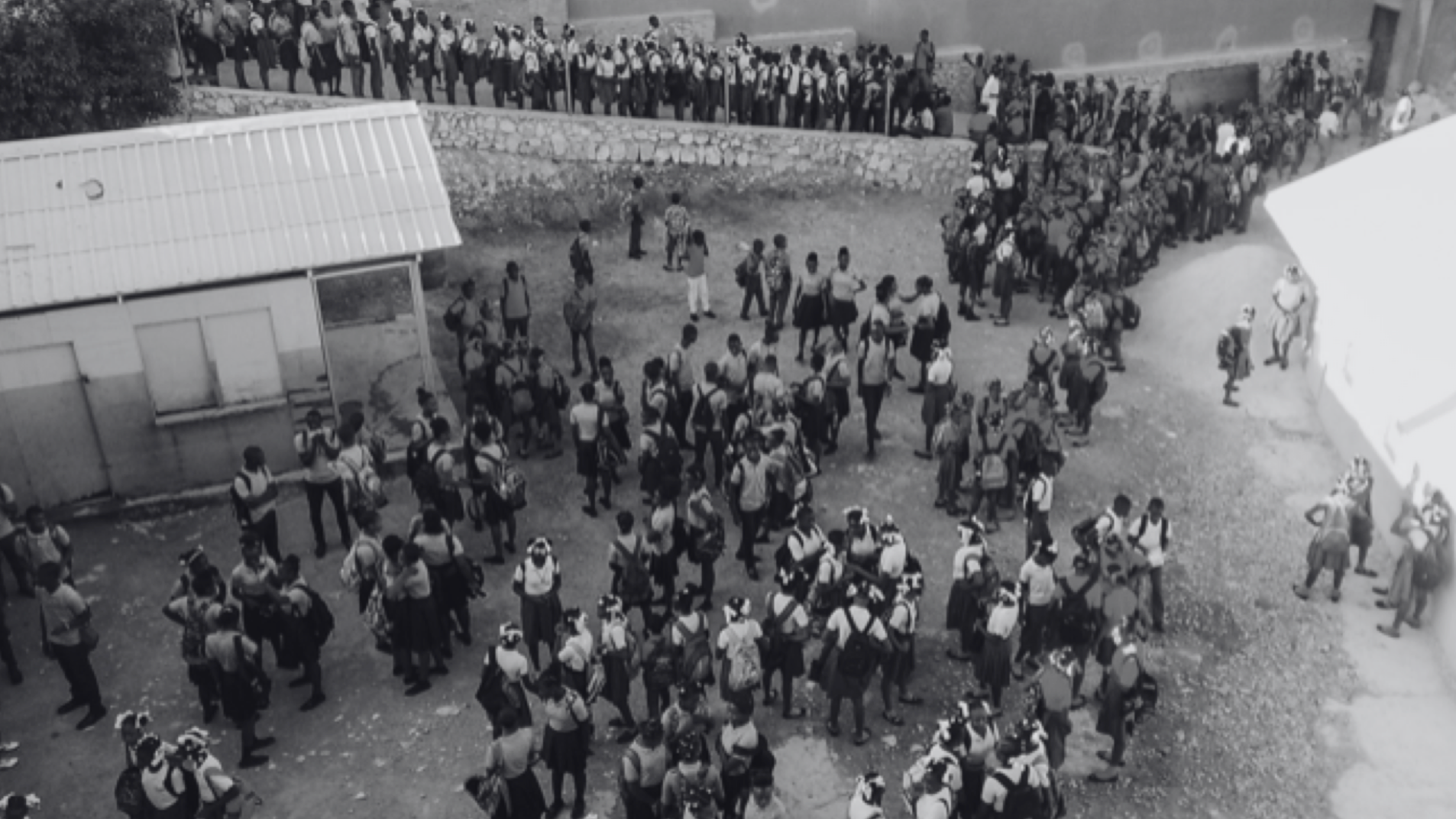 A black and white aerial photograph of a crowded schoolyard filled with students and a few adults. The students, in various uniforms, are grouped in clusters, some standing in lines or forming circles. The schoolyard is surrounded by buildings and has a few trees scattered around. The scene captures a bustling, active environment, possibly during a break or a school event.