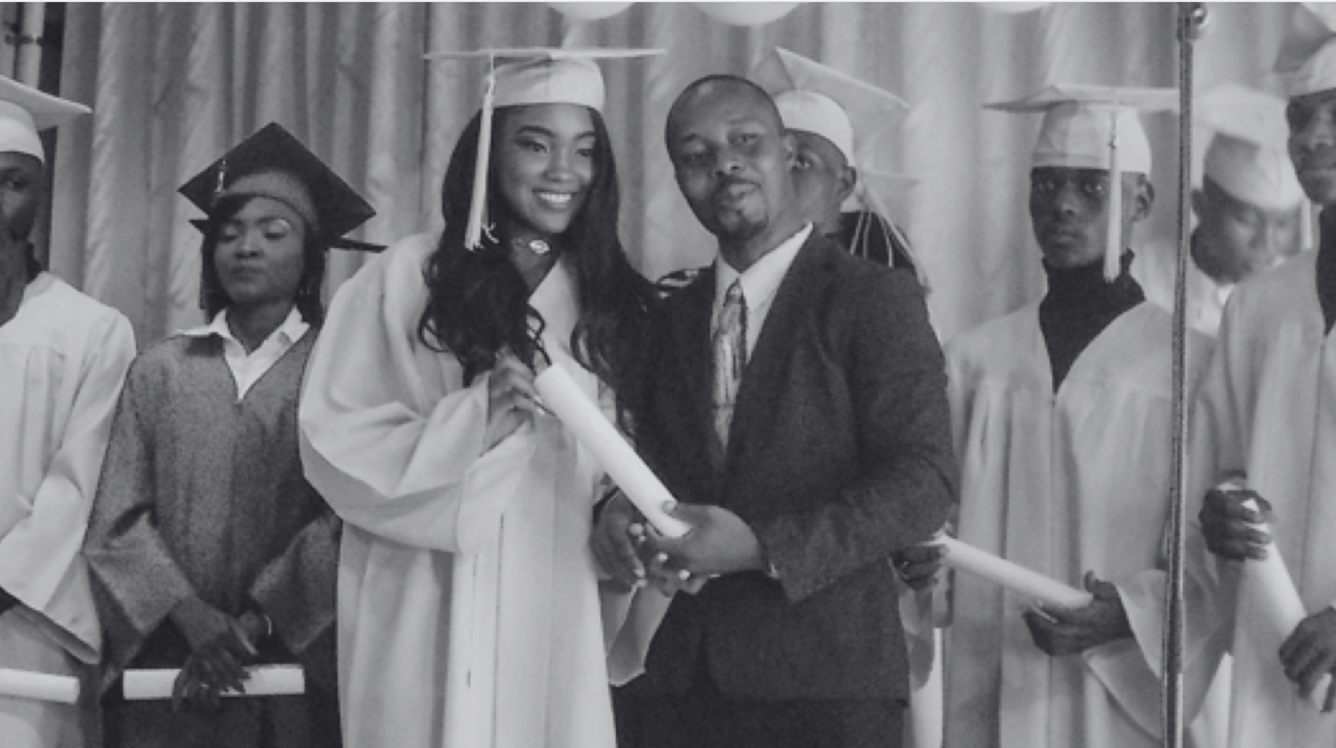 Black and white photo of a graduation ceremony. In the foreground, a proud father, dressed in a dark suit and tie, stands beside his daughter who is in graduation regalia, holding a diploma. She has a broad smile and is looking towards the camera. Behind them, other graduates in white caps and gowns are partially visible, adding to the celebratory atmosphere of the scene.