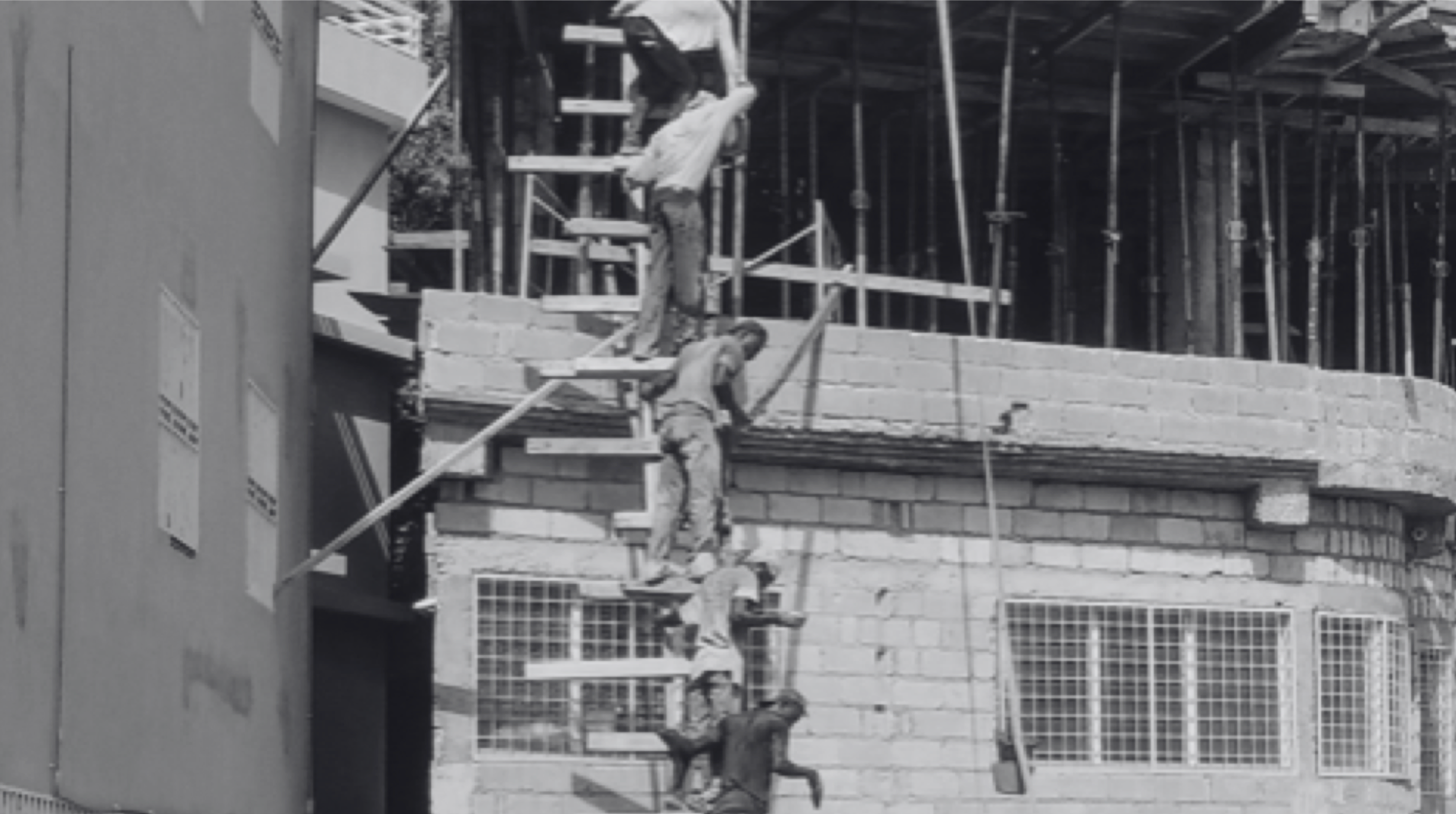 Black and white photo depicting a construction scene where several workers are engaged in building activities. The workers are seen climbing and standing on a makeshift wooden ladder on the exterior of a partially constructed building made of cinder blocks. The image captures the motion and coordination of manual labor in an urban setting.