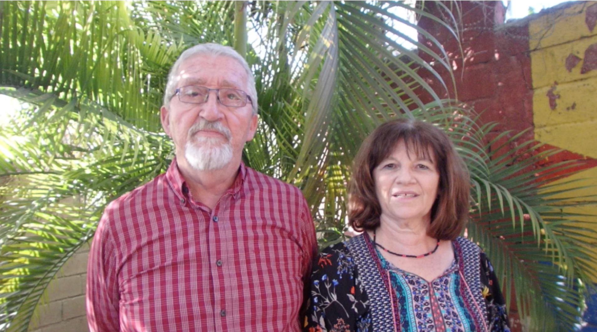 Portrait of an elderly couple standing outdoors with tropical foliage in the background. The man on the left wears glasses and a red checked shirt, and the woman on the right has shoulder-length brown hair and is dressed in a dark floral blouse. Both are smiling slightly and looking directly at the camera, with a rustic, colourful wall partially visible behind them.