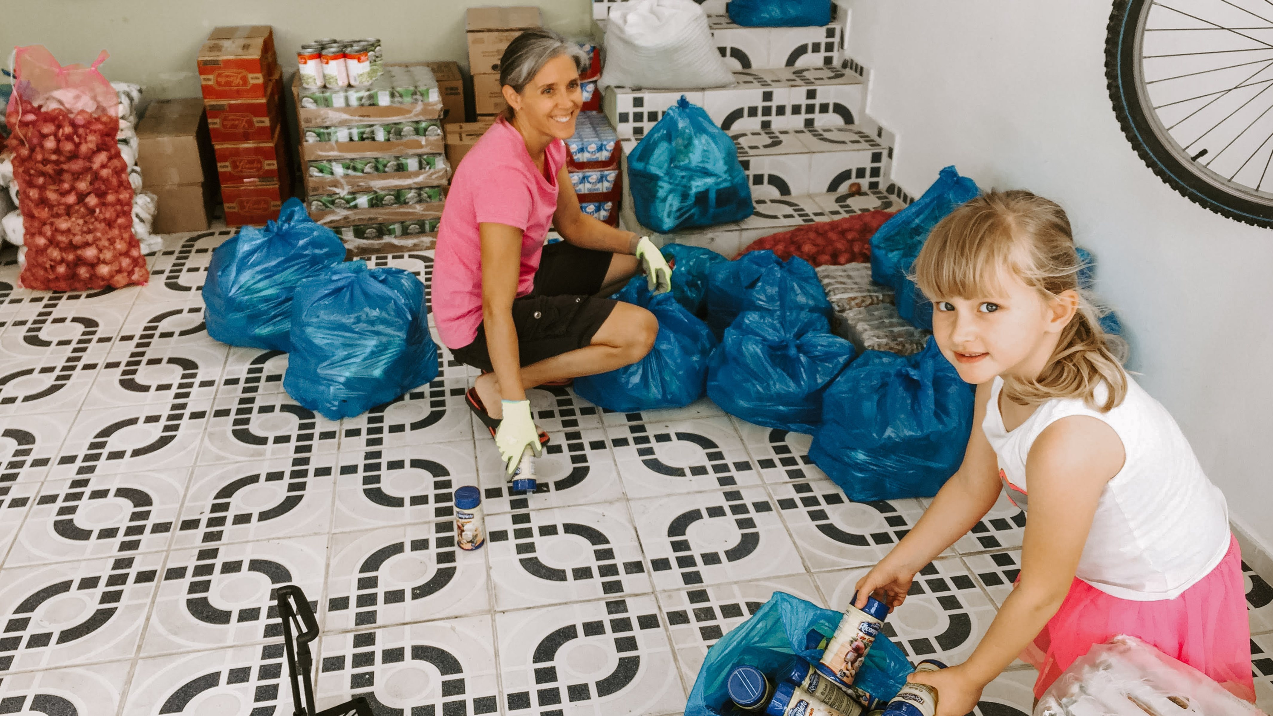 A woman and a young girl, both smiling, sorting food supplies in a spacious room with tiled floors. The woman, in a pink t-shirt and black shorts, is sitting among blue plastic bags filled with groceries, while the young girl, wearing a white tank top and pink skirt, helps organize canned goods. The room also contains other food packages and a bicycle hanging on the wall, indicating a community aid or distribution centre.