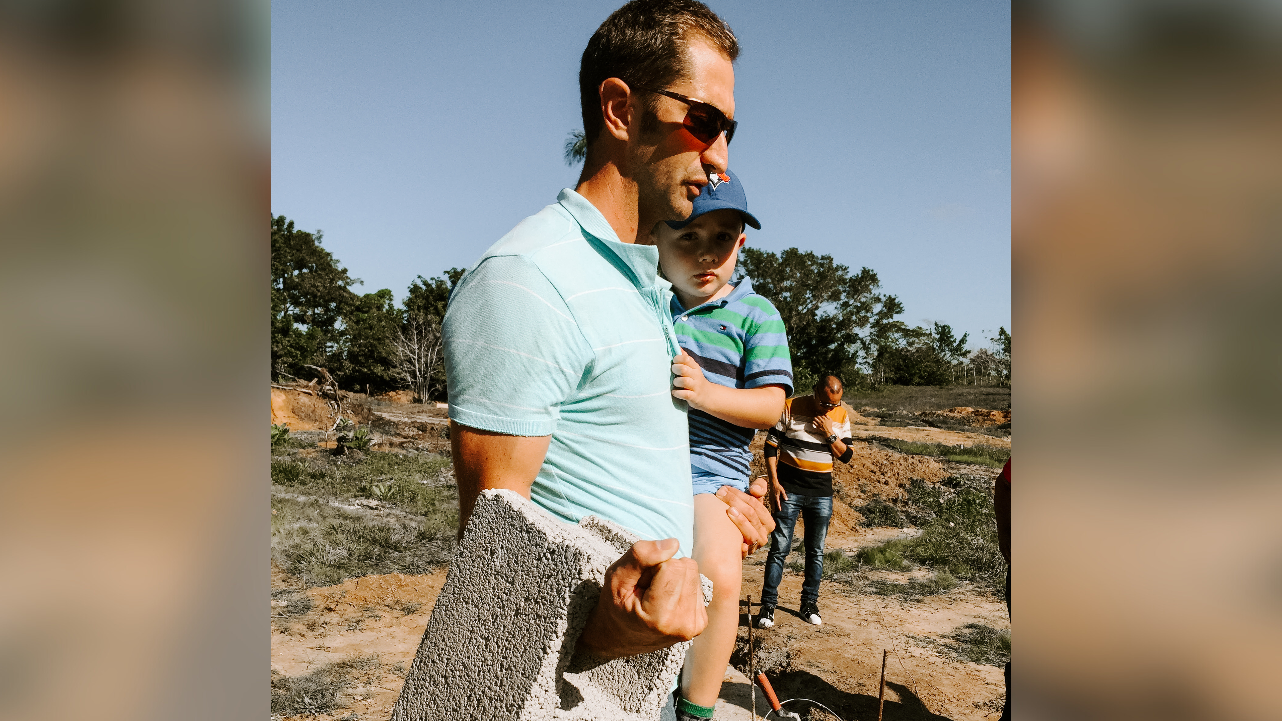 A man in a light teal polo shirt carrying a cinder block with one hand, while holding his young son in the other. The son, wearing a blue and green striped shirt and a baseball cap, appears relaxed. They are outdoors, with construction materials and a couple of workers in the background, suggesting an active building site.