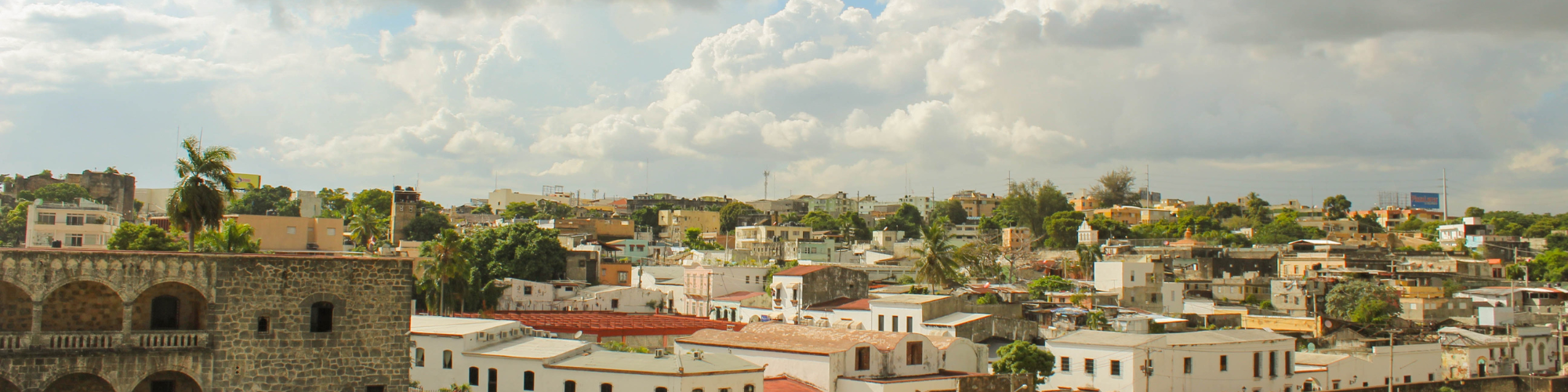 A panoramic view of a city with a mix of historical and modern buildings. The foreground features an old stone building with arched windows, while the background shows a variety of houses and buildings with flat rooftops and colourful facades. The area is dotted with palm trees and other greenery. The sky above is partly cloudy, adding depth to the scene, which captures a vibrant and diverse urban landscape.