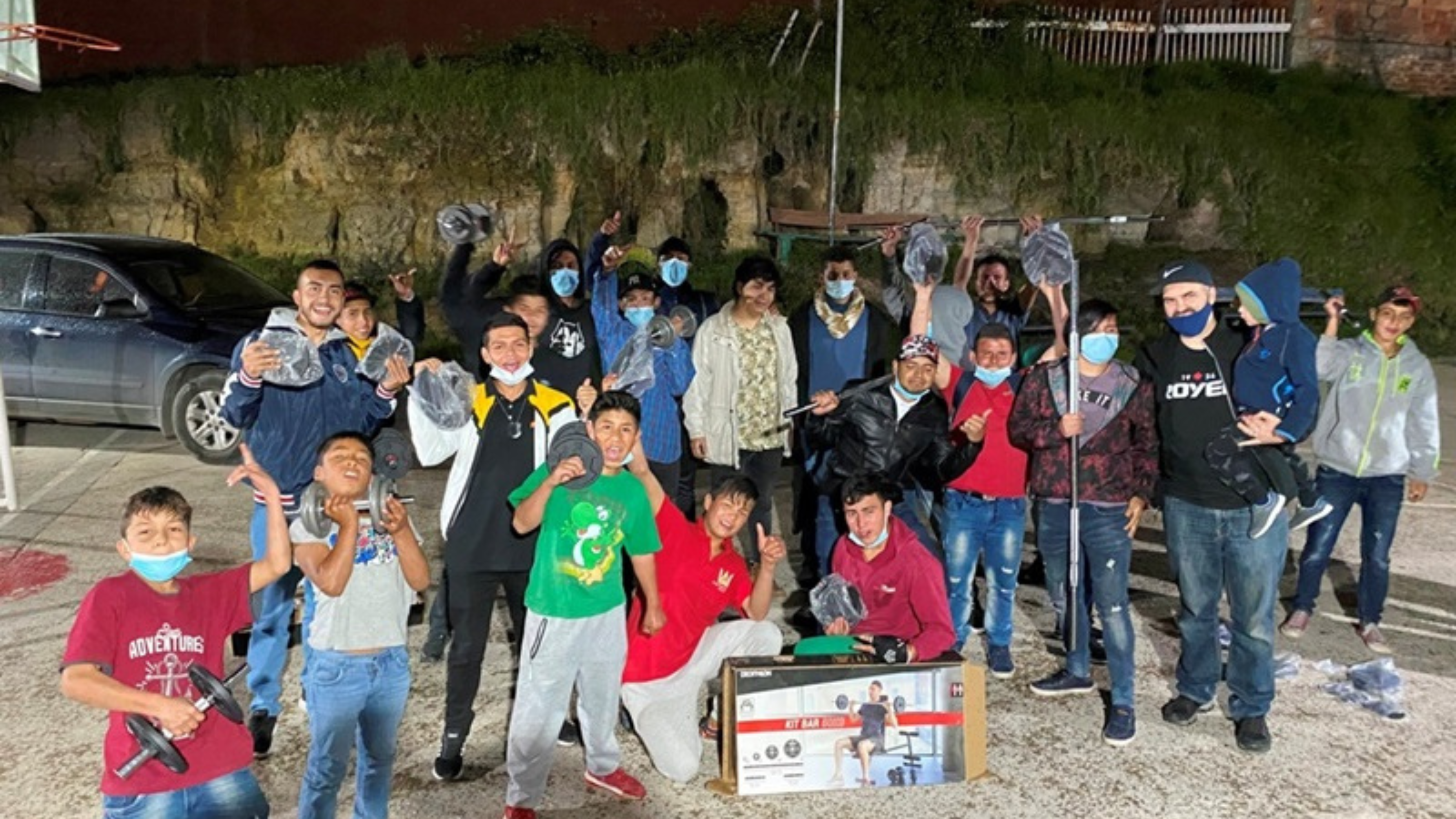 A group of young people, mostly teens, cheerfully pose for a photo at night under streetlights. They are holding up tools and sports equipment, wearing casual clothes and protective face masks. The setting includes parked cars and a rocky hill in the background.